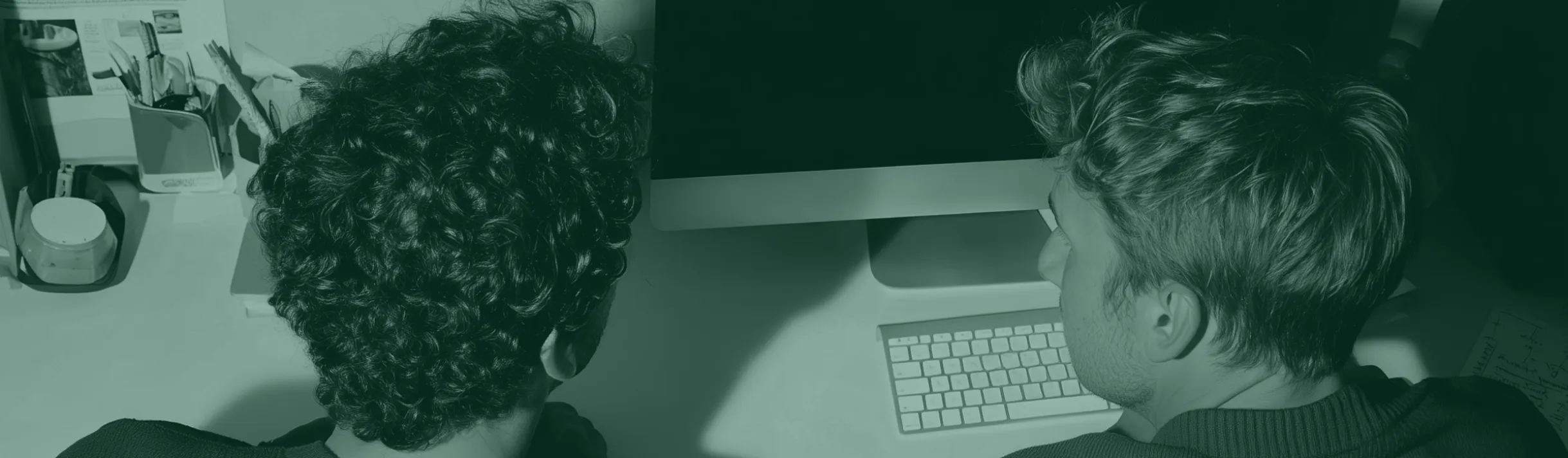 Two people with curly and straight hair working together at a desk with a computer and keyboard.
