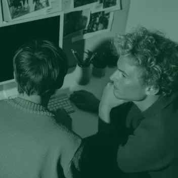 Two people sitting at a desk working together at a computer with photos on the wall behind.