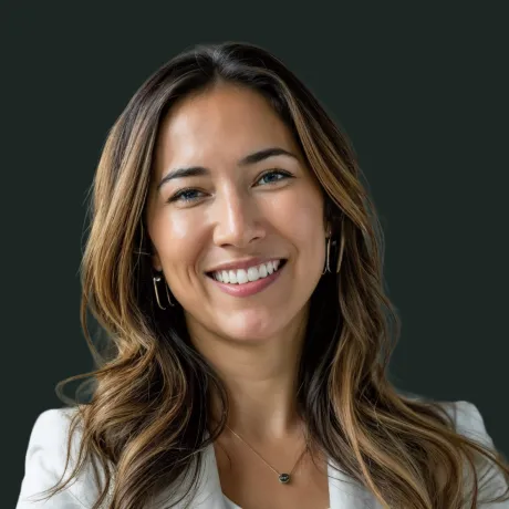 Smiling woman with long wavy hair wearing a white blazer and a pendant necklace against a dark background.