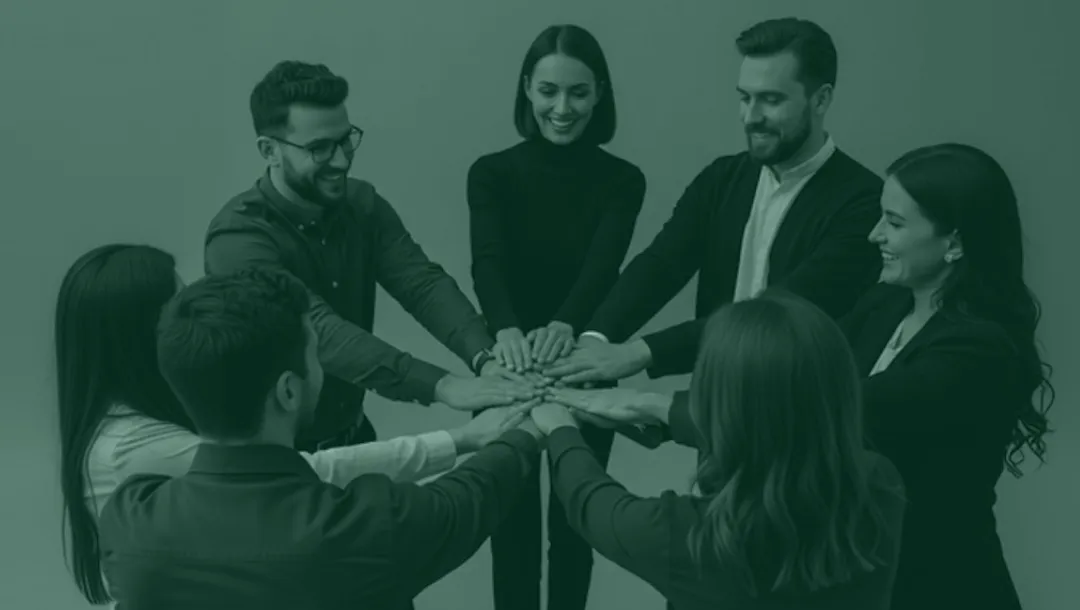 Seven diverse young professionals standing in a circle with their hands stacked together in a show of teamwork and unity.